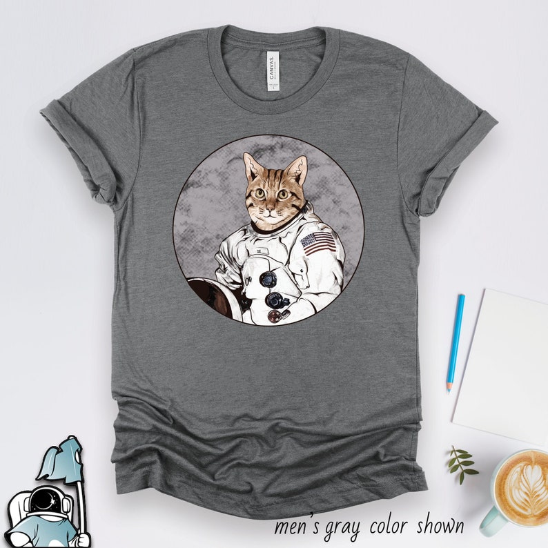 May include: A gray t-shirt with a graphic of a cat wearing an astronaut suit and an American flag patch. The cat is in a circle with a moon-like background.