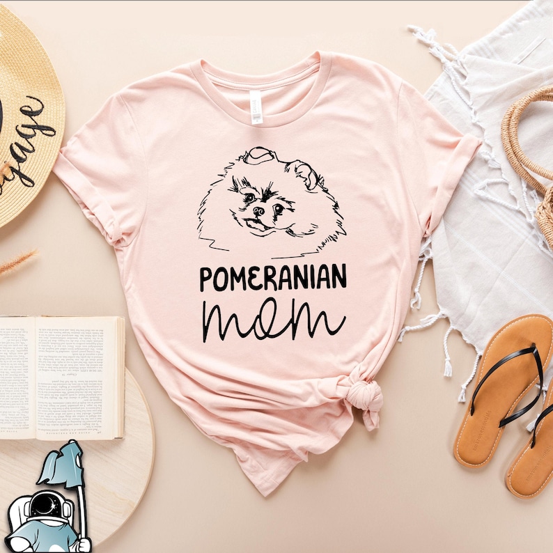 May include: A pink t-shirt with a black line drawing of a Pomeranian dog and the text "Pomeranian mom" printed on the front.