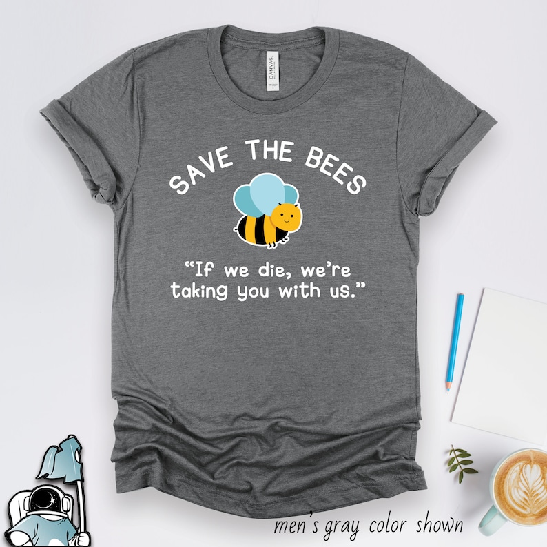 Op de afbeelding: Grijs T-shirt met een witte afbeelding van een cartoon-bij en de tekst "SAVE THE BEES" en "If we die, we're taking you with us."