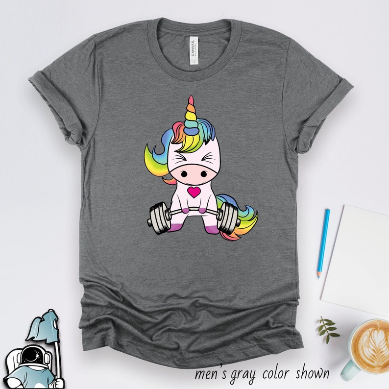 K&ouml;nnte beinhalten: Ein graues T-Shirt mit einem Regenbogen-Einhorn, das Gewichte hebt. Das Einhorn hat ein pinkfarbenes Herz auf der Brust. "Graue Herrenfarbe gezeigt" steht unter dem Bild.
