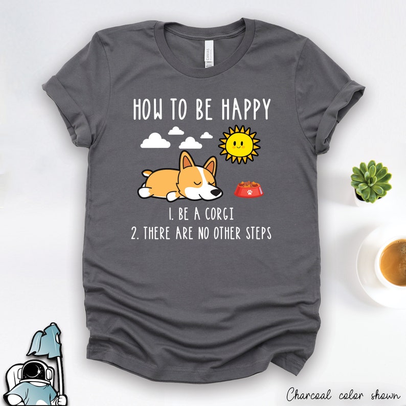 Puede incluir: Una camiseta gris antracita con un dibujo animado de un perro corgi durmiendo en un cuenco de comida. El texto de la camiseta dice "How to be happy", "1. Be a corgi", y "2. There are no other steps".