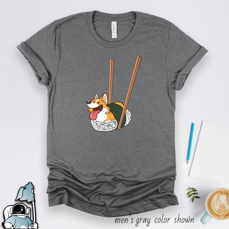 May include: A gray t-shirt with a cartoon image of a corgi dog dressed as a sushi roll. The corgi is being held up by two chopsticks.