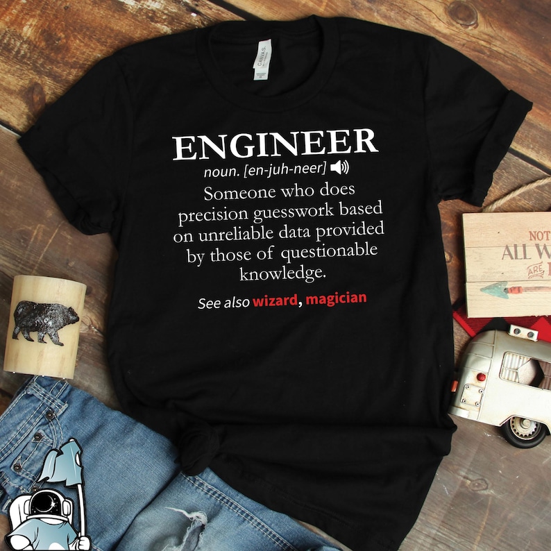 Op de afbeelding: Zwart T-shirt met het woord "ENGINEER" in het wit en de definitie ervan. De definitie beschrijft op humoristische wijze een ingenieur. De zin "See also wizard, magician" is ook opgenomen. Een humoristisch cadeau-idee.