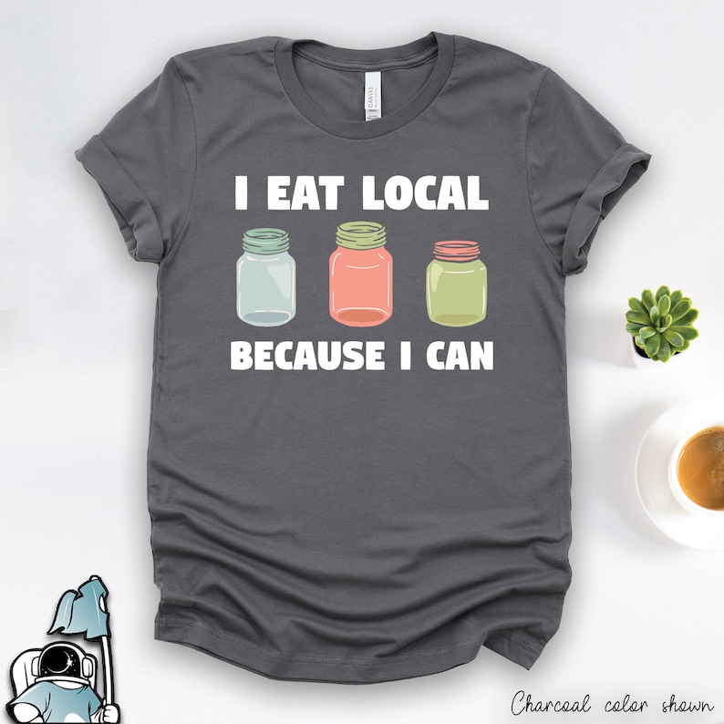 May include: A charcoal gray t-shirt with white text that reads 'I eat local because I can.' Three mason jars with different colored lids are pictured below the text.