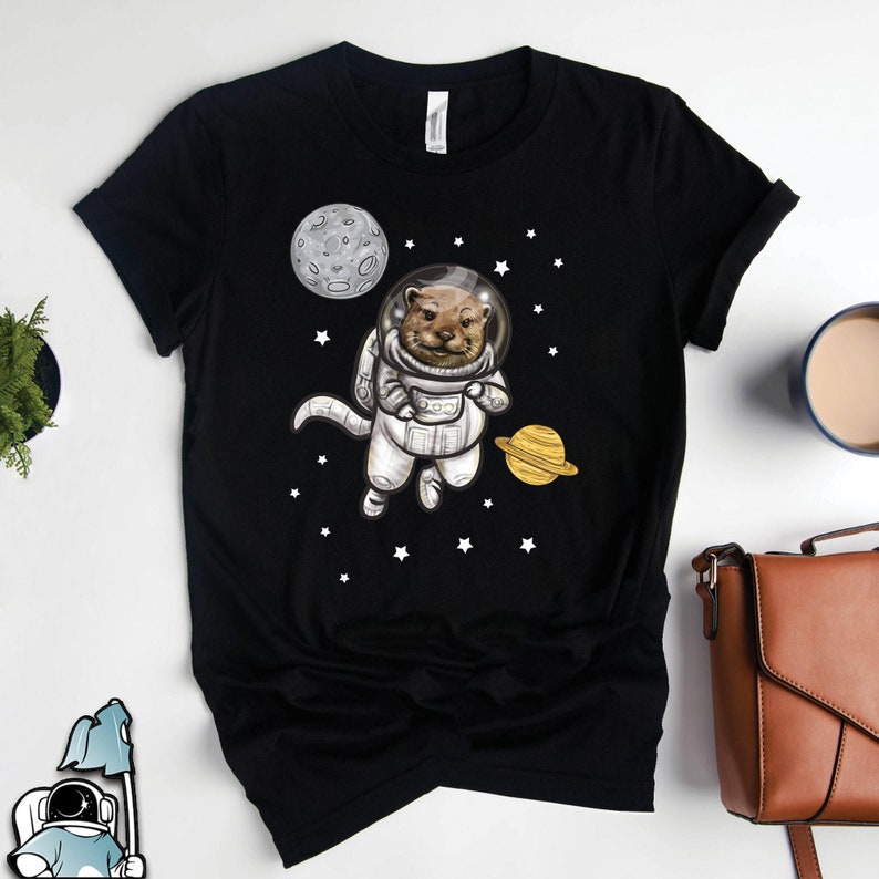 May include: A black t-shirt with a graphic of an otter wearing an astronaut suit in space. The otter is floating in space with a planet and a moon in the background.