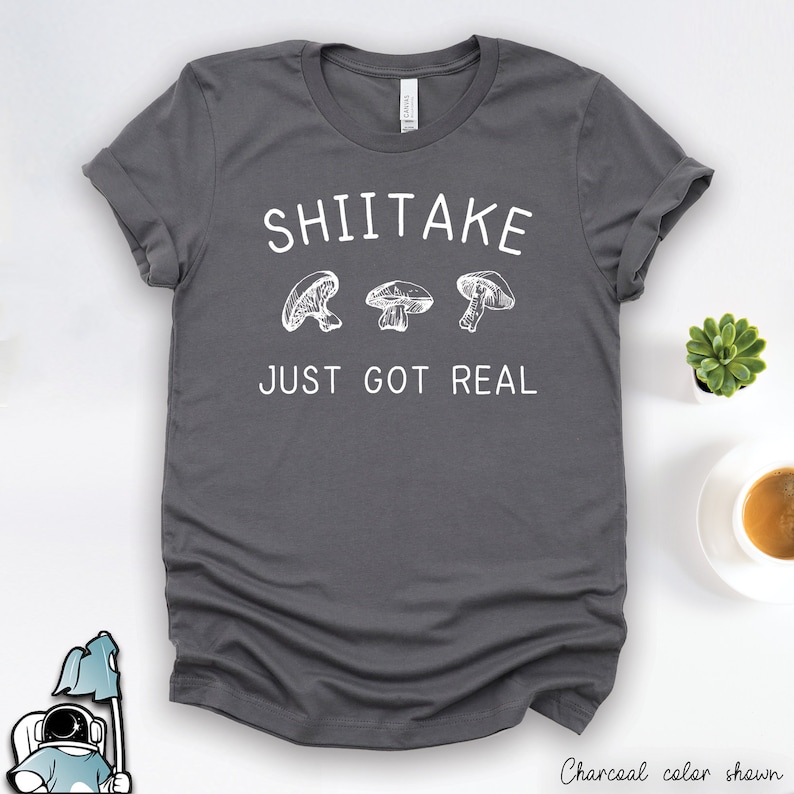 Pu&ograve; includere: Una maglietta grigio antracite con testo bianco che recita "Shiitake Just Got Real" e tre illustrazioni di funghi.