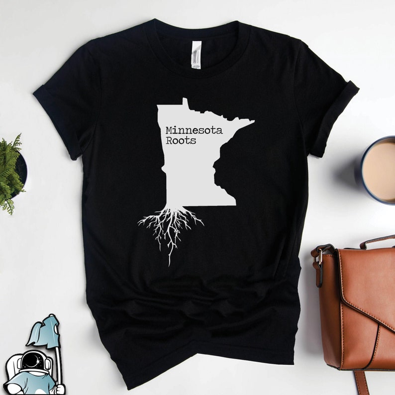 Puede incluir: Camiseta negra con un contorno blanco del estado de Minnesota y el texto "Minnesota Roots" con ra&iacute;ces de &aacute;rbol blancas que se extienden desde la parte inferior del estado.