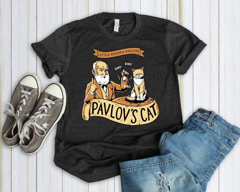 May include: A dark gray t-shirt with a cartoon illustration of a cat and a man with a beard. The text "Little Known Failure!" is above the illustration. The text "Pavlov's Cat" is below the illustration.