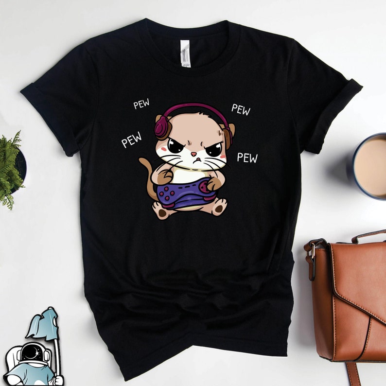 May include: Black t-shirt featuring a cartoon of an angry cat wearing headphones and holding a video game controller. The words 'PEW' are written around the cat. The cat is light brown with dark brown eyes and purple headphones. The t-shirt is a classic crew neck style.