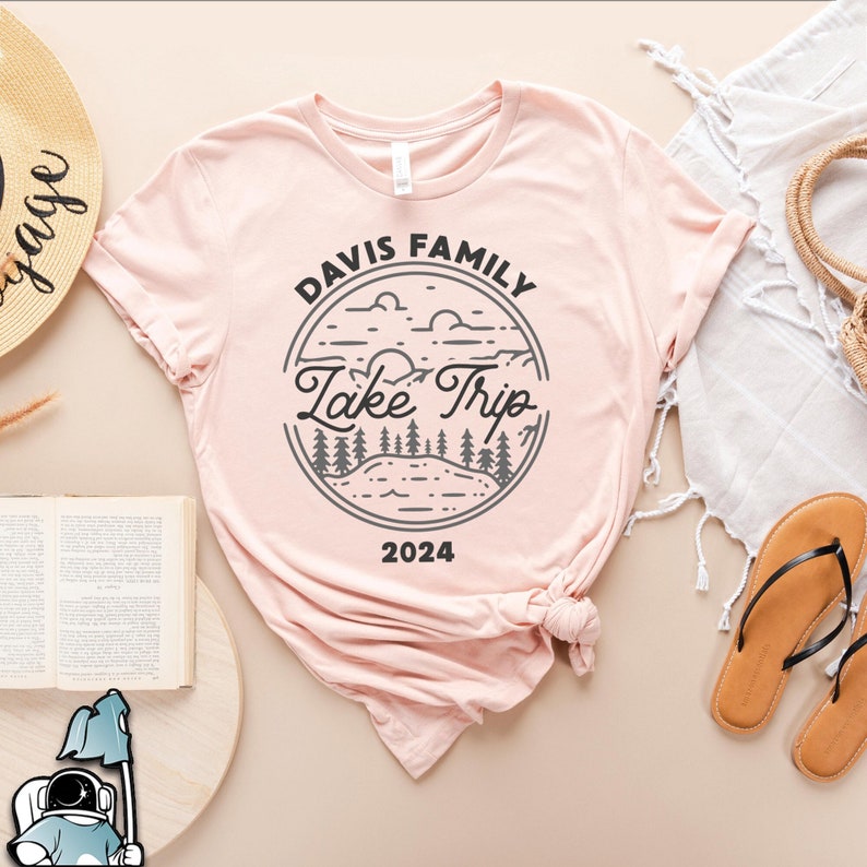 May include: A pink t-shirt with a black and gray graphic design. The design is a circle with the text 'Davis Family Lake Trip 2024' inside. The graphic also includes a stylized image of a lake, trees, and clouds.