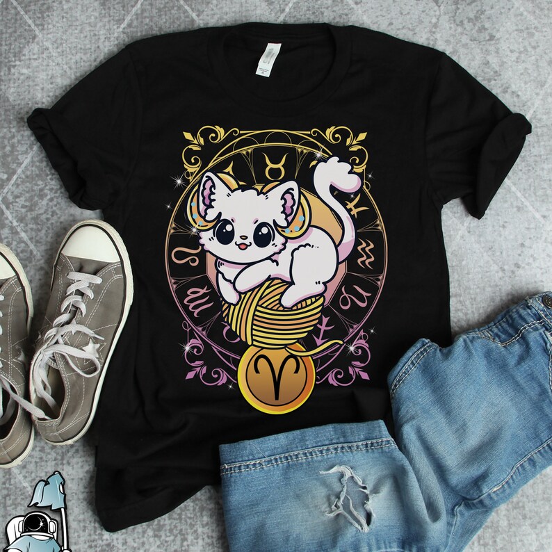 May include: Black t-shirt featuring a white cat holding a ball of yarn. The cat is surrounded by a gold zodiac symbol and the text "Is du" and "U" in a circle.