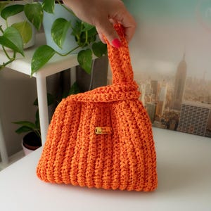 May include: Vibrant orange crocheted handbag with a knotted handle. The bag has a textured, ribbed appearance and a small tag that reads "Nom OnTheRock". The bag is held up by a hand, and the background includes plants and a cityscape.