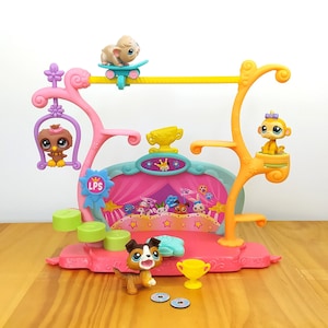 May include: A pink, yellow, and blue plastic playset with a stage, a swing, a skateboard, and a platform. There are four small plastic animal figures on the playset. The playset is on a wooden surface.