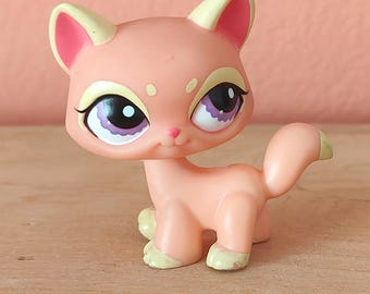 May include: A light pink cat figurine with large purple and black eyes, yellow-tipped ears, and a curled tail. The toy cat has a small pink nose and yellow-green paws. The figurine is on a light brown surface.