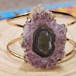 May include: A gold metal bangle bracelet with a large amethyst crystal. The crystal is a cluster of purple crystals with a dark purple center.