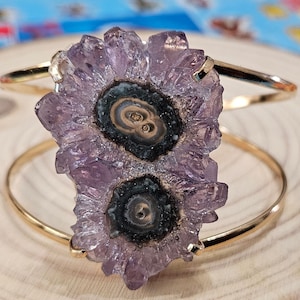 May include: A gold bangle bracelet with a large amethyst crystal cluster featuring two black and white druzy inclusions. The crystal is purple and has a rough, natural texture.