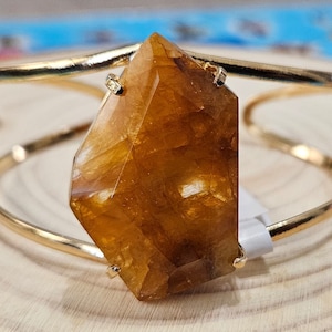 May include: A gold bangle bracelet with a large, faceted, amber-colored gemstone. The gemstone is set in the center of the bracelet and is surrounded by a thin gold band.
