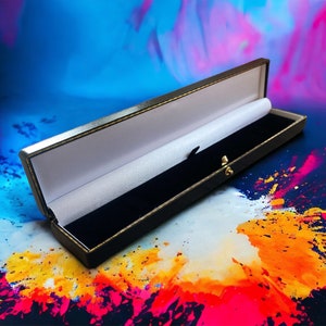 May include: A black velvet-lined jewellery box with a gold clasp. The box is open and the lid is propped up. The box is sitting on a colourful background with splashes of paint.