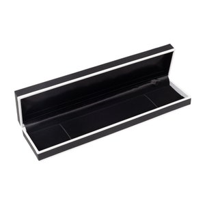May include: A black rectangular box with a white trim and a black velvet interior. The box is open and the lid is propped up.