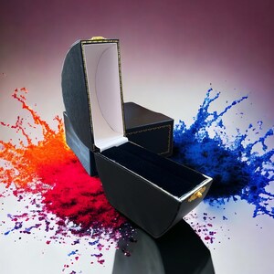 May include: A black ring box with a gold trim and a white velvet interior. The box is open and is sitting on a white surface with red, orange, and blue paint splatters.