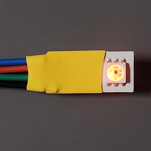 May include: An LED strip with four wires: blue, red, green, and black. The wires connect to a white connector with a lit red LED. The wires are secured with yellow tape. The background is grey. This is a component for electronics.