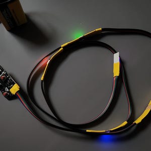May include: An electronic circuit featuring a 9-volt battery, a small circuit board, and black wires. The wires are secured with yellow tape and display red, green, and blue lights.