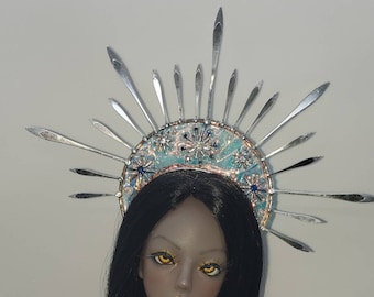 May include: A doll with dark hair and a light brown complexion wearing a white dress with feathers and a silver crown with blue and white accents.