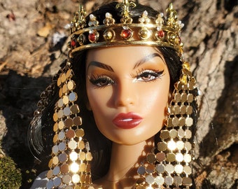 May include: A doll wearing a gold crown with red jewels, a gold chainmail collar, and a blue velvet dress with gold trim. The doll has long black hair and brown eyes.