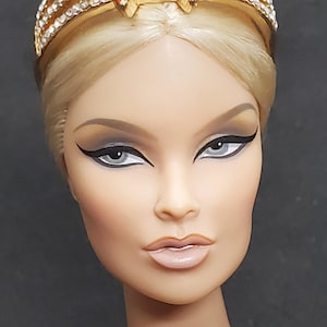 Puede incluir: Cabeza de muñeca rubia con maquillaje dramático, luciendo una corona dorada. La corona presenta acentos de piedras preciosas rojas y brillantes transparentes. La cara de la muñeca es pálida con rasgos definidos y delineador oscuro.