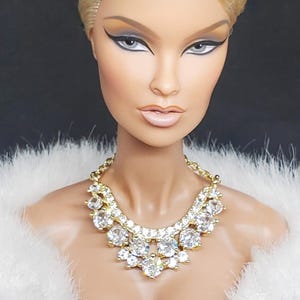 May include: A fashion doll with blonde hair and striking eye makeup, adorned with a gold necklace set with large, clear gemstones. The doll is wearing a white, fluffy boa, against a dark backdrop. The necklace is the main feature.