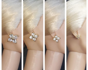 May include: Four pairs of gold earrings with clear crystal stones in a flower shape. The earrings are shown on a doll's ear.