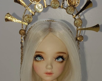 May include: A gold crown with a star-shaped design and clear glass accents. The crown is worn by a doll with white hair and blue eyes.
