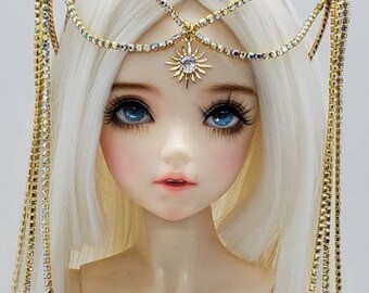 May include: A white doll with blonde hair wearing a gold and crystal crown with dangling chains. The crown features a large star and smaller stars.