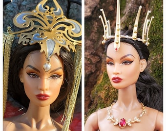 May include: Two fashion dolls with elaborate gold and silver crowns. The doll on the left has a silver crown with a pearl pendant and a gold and silver headpiece with long gold tassels. The doll on the right has a gold crown with a red gem and a gold necklace with a red gem. Both dolls are wearing red dresses.