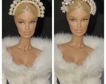 May include: Two doll portraits showcasing elaborate headpieces. The first features a gold-toned tiara with pearls and clear stones. The second displays a similar tiara with floral and clear stone accents. Both dolls wear white, fluffy-textured clothing.