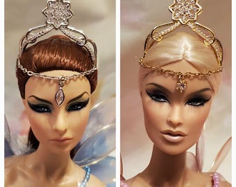 May include: Two dolls with different hair colors wearing elaborate jeweled headbands. The headband on the left doll is silver with clear rhinestones, while the headband on the right doll is gold with clear rhinestones.