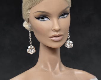 May include: A fashion doll with blonde hair in a low bun. The doll has striking eye makeup and wears silver flower earrings. The doll is dressed in a white gown with a sheer neckline. The background is dark.