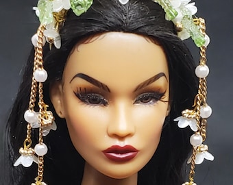 May include: A doll wearing a white floral crown with gold accents and dangling chains. The crown features delicate white flowers, green leaves, and gold beads. The chains are adorned with pearls and small white flowers. The doll has dark hair and red lipstick.