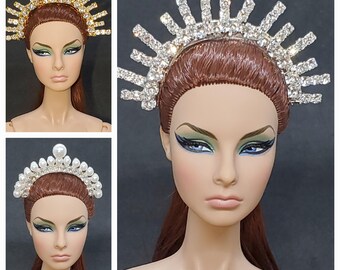 May include: Three tiaras are shown. One is gold with crystal accents, another is silver with clear crystals, and the third is white with pearls. All are set on a doll's head with brown hair, blue eyes, and green eyeshadow.