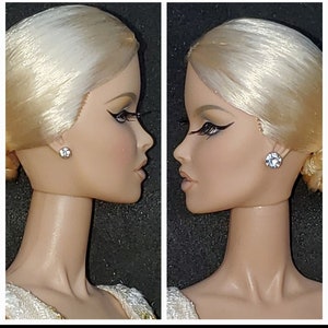 Puede incluir: Primer plano de la cara de una muñeca de porcelana con cabello rubio, que lleva un par de pendientes de cristal transparente. La muñeca mira hacia la derecha.