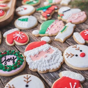 Op de afbeelding: Een assortiment kerstsuikerkoekjes versierd met verschillende ontwerpen, waaronder een sneeuwpop, de kerstman, een pinguin, een want, een krans met het woord "vreugde" en een elf. De koekjes zijn gerangschikt op een houten oppervlak.