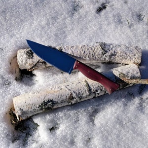 May include: A blue steel knife with a dark wood handle resting on two birch logs in the snow.