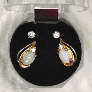 May include: A pair of gold earrings with white opal stones and clear cubic zirconia accents. The earrings are displayed in a black velvet box.