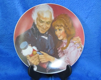 May include: A vintage porcelain plate with a colorful illustration of a man and a girl holding a nutcracker. The plate has a red background and a gold trim.
