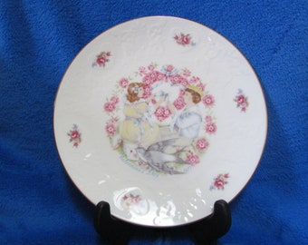 May include: A white porcelain plate with a gold rim features a painted scene of two children exchanging a dove. The plate is decorated with pink and white flowers and the text "Valentine's Day 1977".