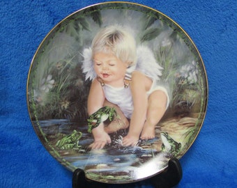 May include: A porcelain plate with a gold rim features a painting of a cherub with wings, playing with frogs in a pond. The cherub is wearing a white dress and has blonde hair. The plate is on a black stand.