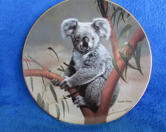 May include: A decorative ceramic plate featuring a detailed painting of a koala bear sitting on a eucalyptus branch. The plate has a gold rim and the artist's signature, "Charles Fracel", is visible in the bottom right corner.
