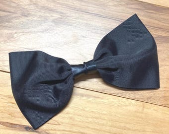 May include: A large, black bow hair accessory. The bow is made of a ribbed fabric and has a leather-like band in the center. The bow is set against a wooden background.