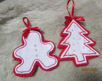 May include: Two felt Christmas ornaments, one shaped like a gingerbread person and the other like a Christmas tree. Both ornaments are white with red outlines and have white pearl beads sewn on.