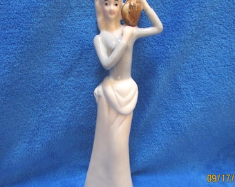 May include: A white porcelain figurine of a woman in a long dress, holding a brown pitcher to her shoulder.